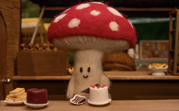 A felt mushroom character behind a bakery counter of tiny cakes